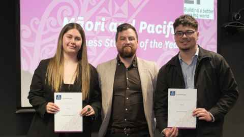 Two students standing in front of a pink backdrop holding certificates, with their teacher.