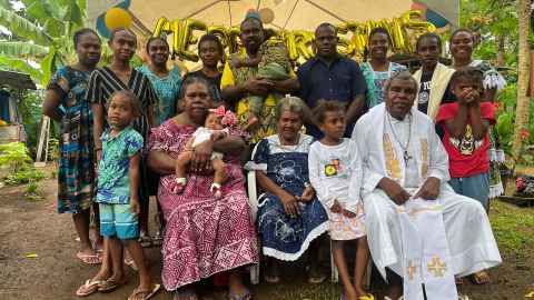 An intergenerational group standing and seated outside, surrounded by foliage and under a cloth gazebo