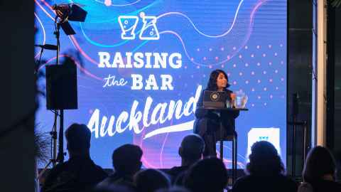 Speaker seated on stage with a laptop and water jug on a table in front of them.