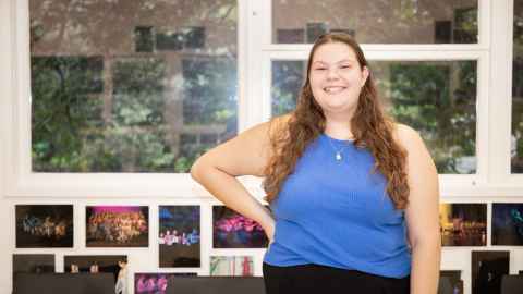 Michaela stands in a dance studio with one hand on her hip and smiles broadly at the camera 