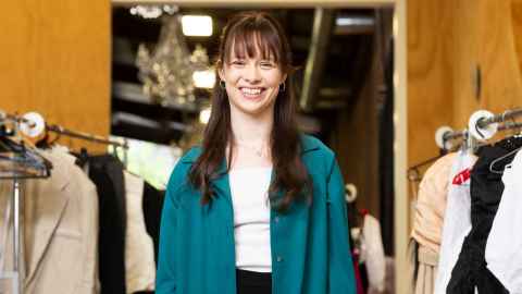 Emma stands amongst costumes backstage and smiles to camera. She wears a green jacket and white tee shirt.