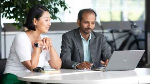 A woman and a man sitting at a table looking at a laptop. The woman is wearing a white shirt and a green skirt, and the man is wearing a grey suit jacket and a light blue shirt.