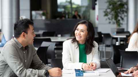 A woman and a man sitting at a table in discussion with each other. They are sitting in a corporate environement. The man is wearing a grey shirt and the woman is wearing a white blazer and a green shirt.