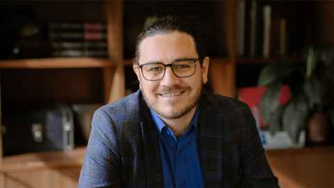 Image of a man with dark hair pulled back into a ponytail, wearing glasses, a dark blazer and a blue shirt, smiling.