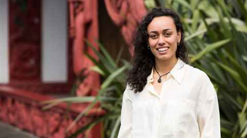 Image of a woman with long brown hair, wearing a white shirt, standing in front of a marae, smiling.