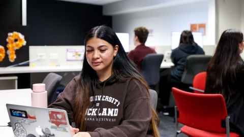 Image of a woman with long brown hair, wearing a brown sweatshirt, sitting at a table working on a laptop.