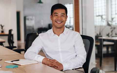 Image of a man with short, dark hair, wearing a while shirt, sitting at a long desk, smiling.