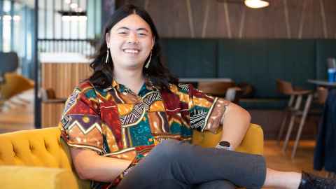 Image of a man sitting on a mustard coloured couch with a colourful shirt on, grey trousers, long dark, smiling.hair, 