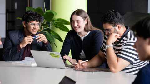 Group of students sitting at a table, looking at a laptop, smiling.