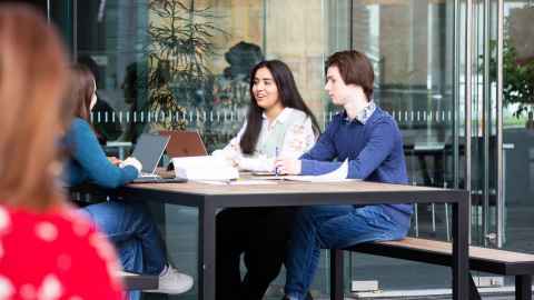 Group of three students sitting outside at a table with laptops and notebooks, talking.