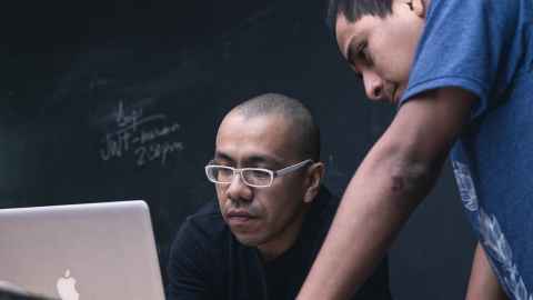 Two men looking at a laptop screen with a chalkboard behind them