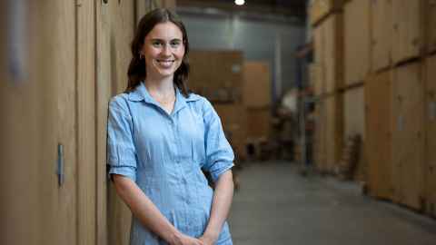 A woman with long brown hair, wearing a light blue dress, leaning against a wall in a warehouse environment.