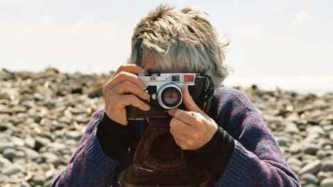 Artist Fiona Clark holds a camera to her face while on a beach. Her arms, head and hands are visible. Her face is obscured by the camera.