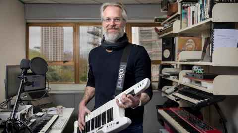 Godfrey De Grut standing in his office with his keytar  (shaped like a guitar, but with a short keyboard where the main body is, and no strings)