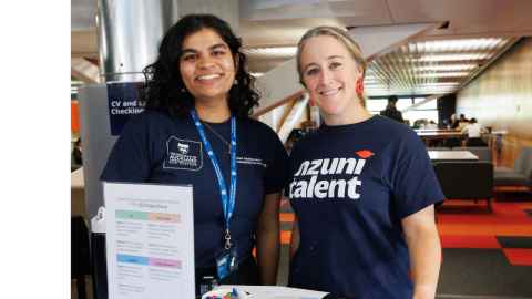 Two female careers adviser wearing blue t shirts smiling to camera, orange and grey carpet and grey sofas in the background