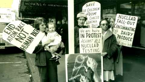 1960s photo of four women, one holding a baby, in front of shops, holding signs protesting against nuclear testing. MSS & Archives A-244.