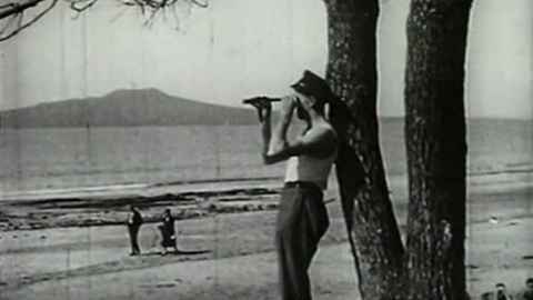 Black and white photo of a person holding a telescope looking out to sea with a beach and rangitoto in the background.