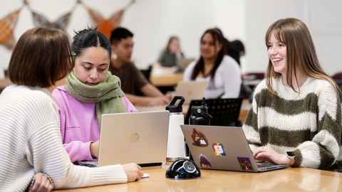 Three female students chatting at a desk with two open laptops on the desk