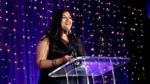 A smiling woman at a microphone with glittering lights behind her.