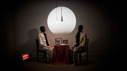 Photograph of two masked figures sitting either side of a small table in a dark room with a moon-like light on the wall