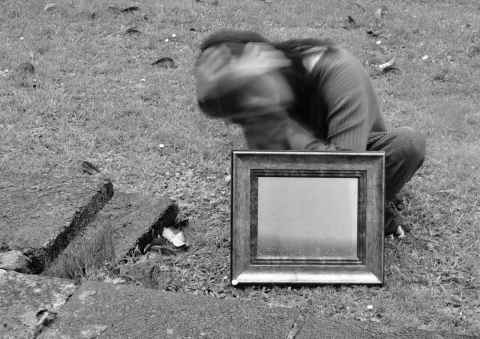 Black and white photograph of blurred person crouched behind a framed small mirror.