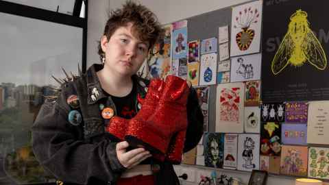 Kai stands in the bedroom wearing a spiked jean jacket with pins and hold glittery red platform text. 
