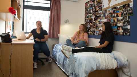 Three students in a private room, one is by the desk and two are on the bed