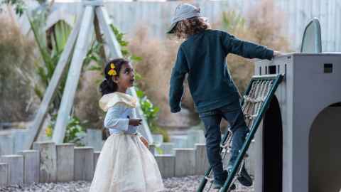 Two children, one stands on a playground set looking down at the other dressed in a princess costume.