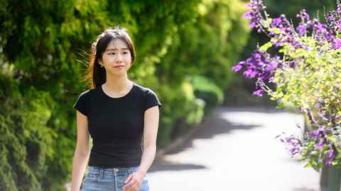 Woman in black t-shirt and jeans, walking down a path with some purple flowers on the right