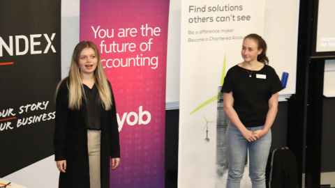 Two women standing at the front of a lecture theatre, speaking to an audience. They are standing infront of banners that have information about accounting on them.