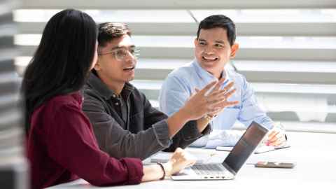 Three students sitting at a table in a light-filled room in conversation with each other.